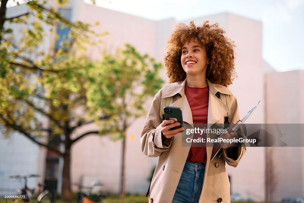 Smiling student walking outside university building using smartphone