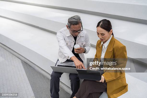 two senior business professionals, collaborating outdoors, working together on their laptop - voorzitter stockfoto's en -beelden
