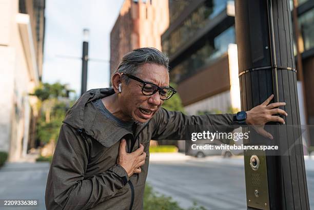 senior man suddenly feeling a sharp pain in his chest while running, griping a nearby pole for support, looking at his smartwatch to check his condition - panting stock pictures, royalty-free photos & images