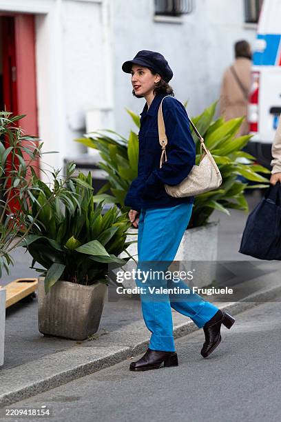 Passerby wears a blue hat, a bue cargo jacket, a beige bag, brown tights, light blue trousers and brown leather heeled mules during the Milan Design...