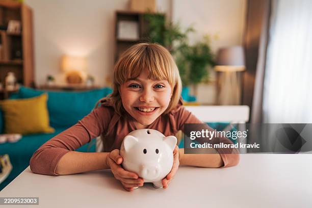 lächelndes mädchen, das zu hause ein sparschwein hält und über ersparnisse lernt - spardose stock-fotos und bilder