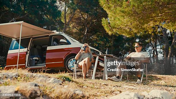 happy mature couple enjoying morning coffee at seaside camping spot with retro camper van - van life stock pictures, royalty-free photos & images