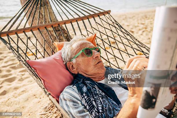 senior man solving puzzle in newspaper while lying down on hammock at beach during vacation - pension de retraite photos et images de collection