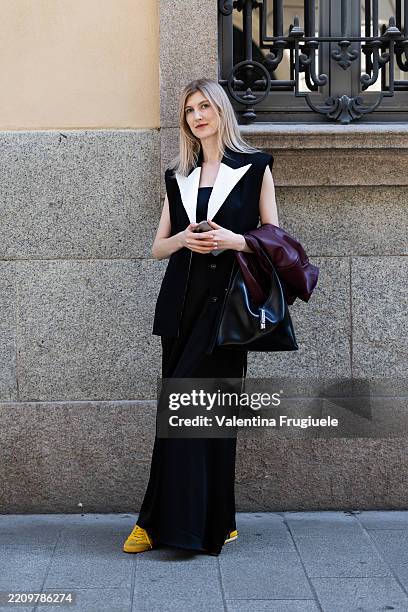 Passerby wears a bordeaux leather jacket, a black leather bag, yellow sneakers, a black long sleeveless dress and a black and white gilet during the...