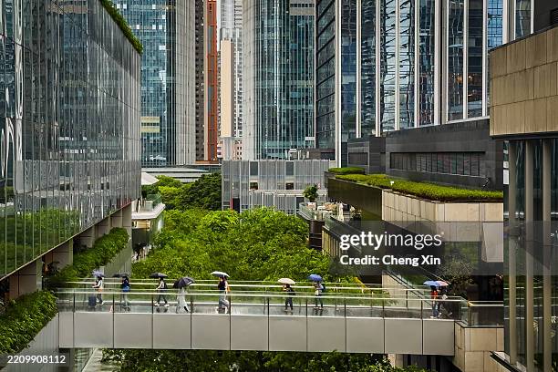People holding umbrellas walk across a skybridge connecting two sections of a modern shopping center surrounded by high-rise office buildings and...