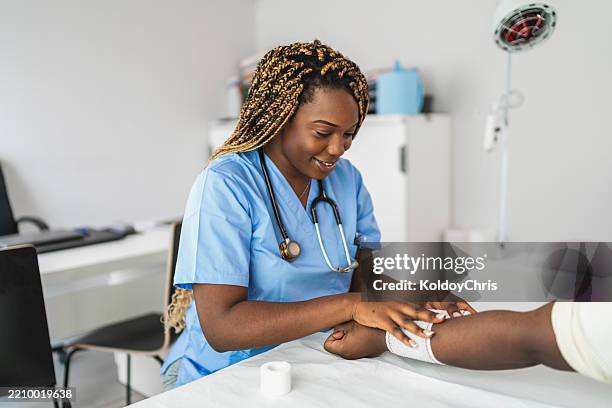smiling african american doctor bandaging patient's arm in medical office - wundversorgung stock-fotos und bilder