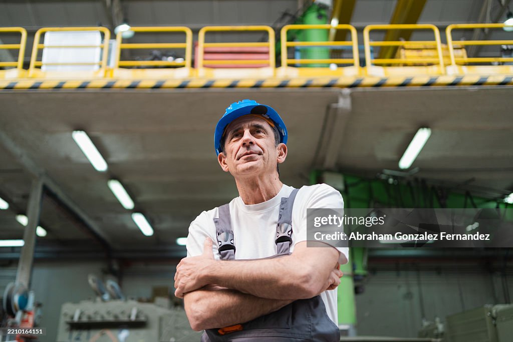Senior factory worker standing confidently, arms crossed, amid industrial machinery, gazing upward