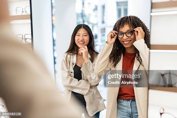 two young women trying eyeglasses in optical store smiling at mirror - provare foto e immagini stock