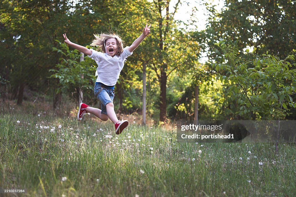 Glückliches kleines Kind trägt ein weißes Hemd und rote Turnschuhe, die an einem heißen und sonnigen Sommer- oder Frühlingstag auf einer Wiese springen