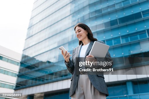 Smiling businesswoman using smartphone and holding laptop in front of office building Smiling businesswoman using smartphone and holding laptop in front of office building