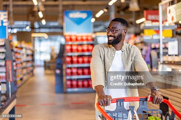lächelnder mann, der in einem modernen supermarkt mit einem schubkarren einkauft - supermarkt gang stock-fotos und bilder