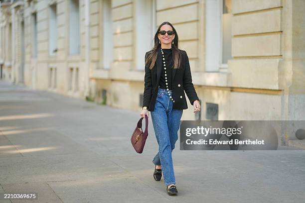 Alba Garavito Torre wears long straight brown hair styled with a center part. Black rectangular sunglasses are styled with large gold geometric...