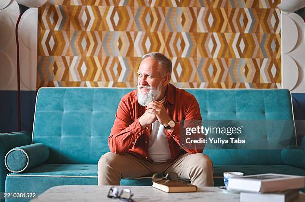 älterer mann sitzt auf dem sofa und schaut weg, während er über etwas nachdenkt - vorruhestand stock-fotos und bilder