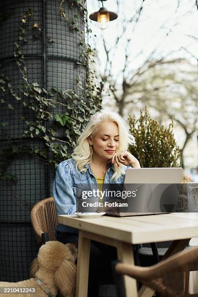 photo of a beautiful woman in her fifties, sitting in the garden of a cafe and working on a laptop computer. - laptop cafe stock pictures, royalty-free photos & images