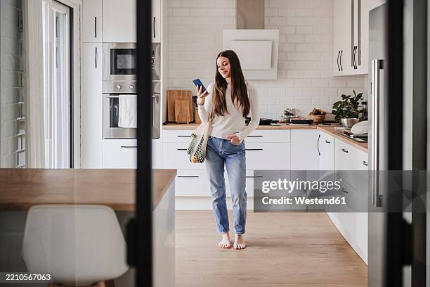 smiling woman in kitchen using mobile phone holding mesh bag with vegetables - barefoot stock pictures, royalty-free photos & images