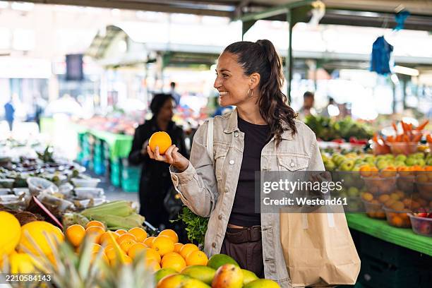 woman shopping at a local market holding an orange and a paper bag - choosing stock pictures, royalty-free photos & images