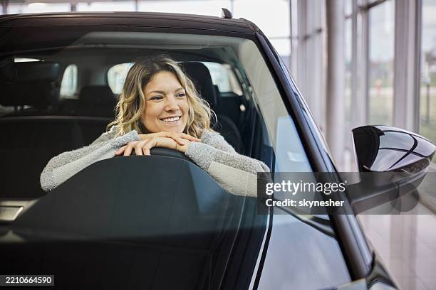 mujer feliz disfrutando dentro de un coche en una sala de exposición. - mujeres de mediana edad fotografías e imágenes de stock