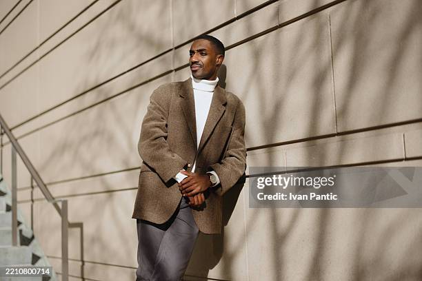 confident man relaxing outdoors in stylish winter fashion against a minimalist wall - black blazer stock pictures, royalty-free photos & images