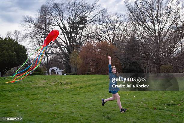 flying kite with colorful streamers. 11-year-old girl runs across park meadow during cloudy day. - blue dress stock pictures, royalty-free photos & images