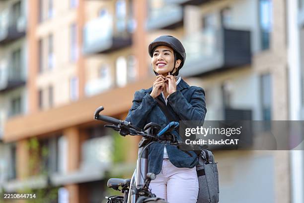 mujer sonriente sujetando casco de bicicleta al aire libre cerca de edificios de apartamentos urbanos - autoconsumo electrico fotografías e imágenes de stock