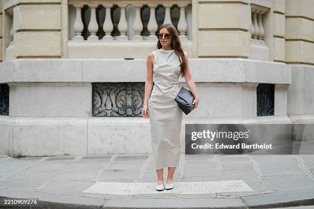 Diane Batoukina wears an off-white sleeveless midi dress by St. Agni featuring an asymmetrical pleated structure at the waist and a high neckline. A...
