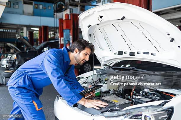 ingeniero mecánico, hombre de uniforme, trabaja con un coche eléctrico roto. .capó de un coche eléctrico mientras se trabaja en un taller. concepción del servicio de coches. fijación de vehículo eléctrico. ev. mecánico revisando el voltaje de la b - pieza de máquina fotografías e imágenes de stock