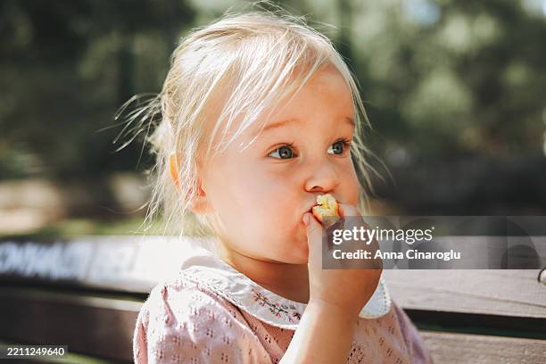 an adorable little girl eats a snack outside, looking content and happy in the sunlight. - weibliches baby stock-fotos und bilder