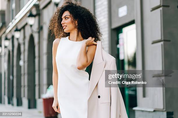 smiling beautiful mixed race woman walking in the city and holding her jacket - white dress stock pictures, royalty-free photos & images