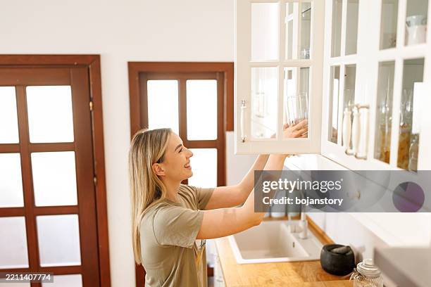 young woman taking drinking glasses from a kitchen cabinet - armário imagens e fotografias de stock
