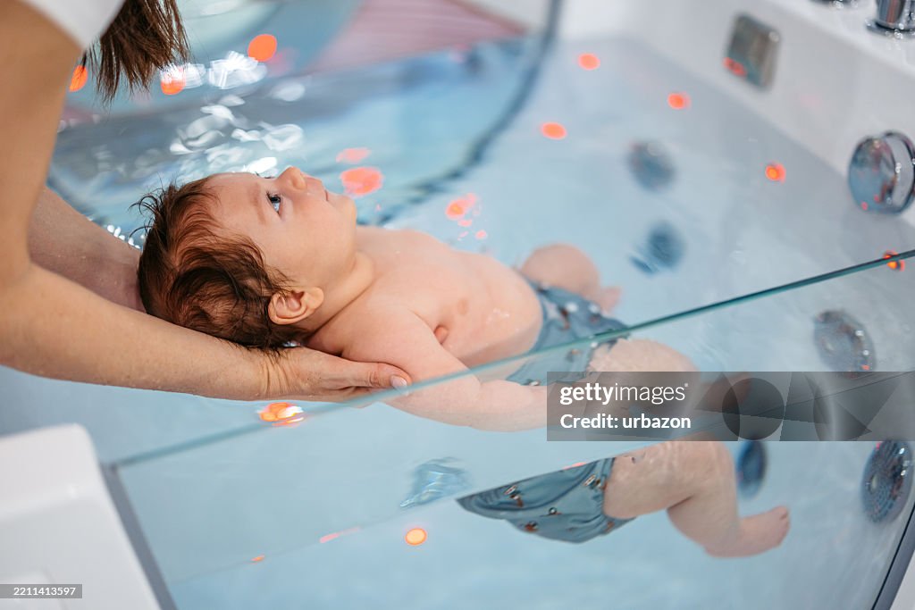 Mutter im mittleren Erwachsenenalter, die ihren kleinen Jungen hält, während er in einer Wanne im Baby Spa schwimmt