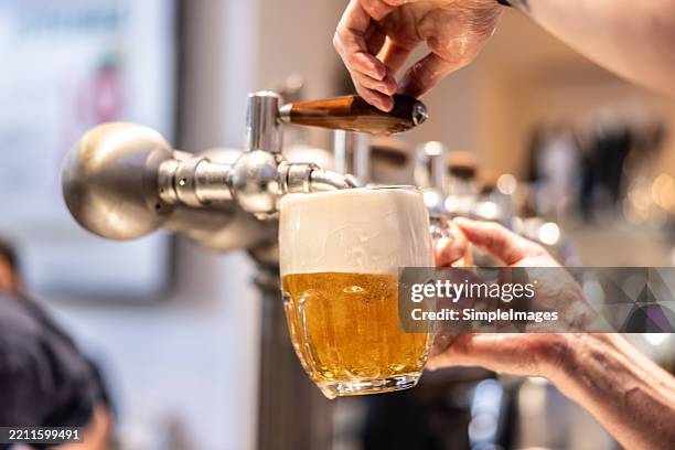 a close-up of an unrecognisable person's hand holding a pint glass at an angle being filled with lager. the lager is golden in colour creating foam and bubbles. - bierfest stockfoto's en -beelden