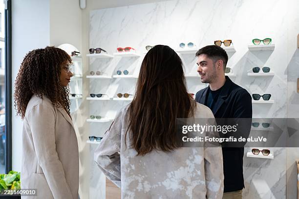 optician helping customers choosing eyeglasses in modern optical store - occhiali e lenti foto e immagini stock