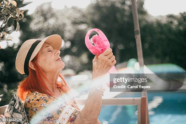 senior woman in floral summer dress enjoys cool air from electric plastic pink fun in summer afternoon - hand fan stock pictures, royalty-free photos & images