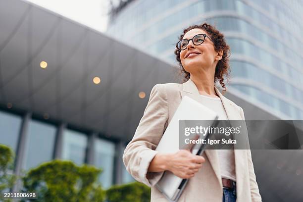 smiling businesswoman holding laptop and looking away near office building - motivatie stockfoto's en -beelden