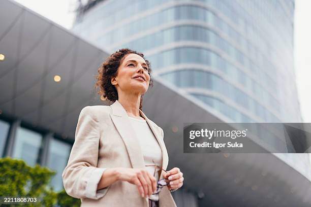 confident businesswoman holding eyeglasses and looking away near modern office building - vision bildbanksfoton och bilder