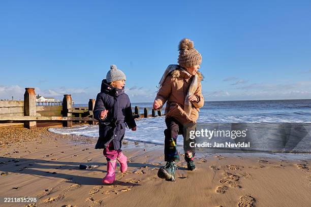 two sisters running along winter beach – joyful childhood adventure by the sea - real family stock pictures, royalty-free photos & images
