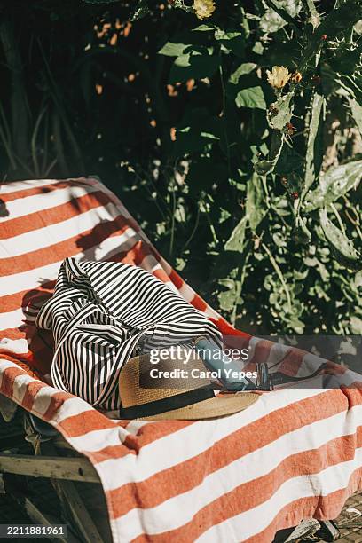 sun lotion, straw hat, summer bag rest on a striped towel outdoors, warm summer light - striped towel stock pictures, royalty-free photos & images