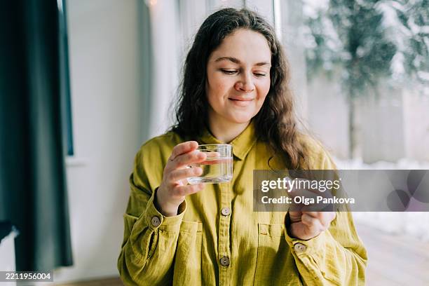 smiling woman holding pill and glass of water, taking medicine at home - vitamina fotografías e imágenes de stock