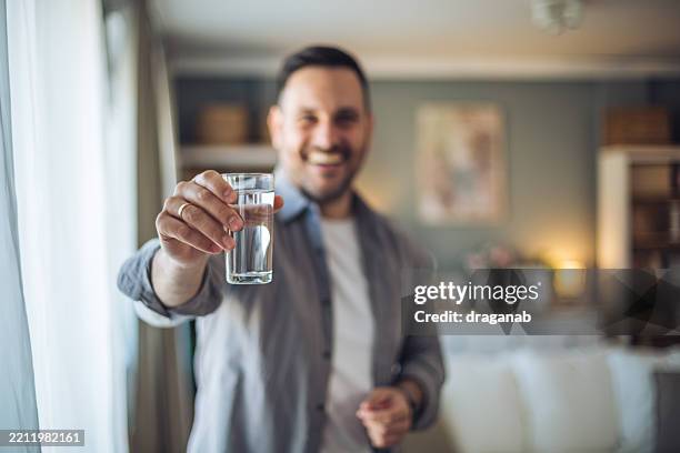 lächelnder mann zeigt glas wasser zu hause - destilliertes wasser stock-fotos und bilder