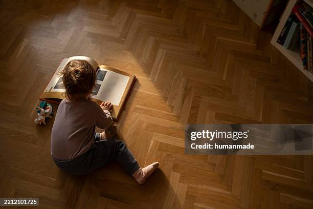 baby girl sitting on hardwood floor reading a book in natural sunlight - houten vloer stockfoto's en -beelden