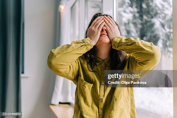 woman covering her eyes with her hands by the window, expressing sadness and despair - winterdepression stock-fotos und bilder