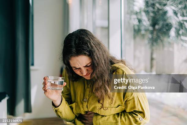 woman suffering from stomach ache holding glass of water by window - lebensmittelvergiftung stock-fotos und bilder