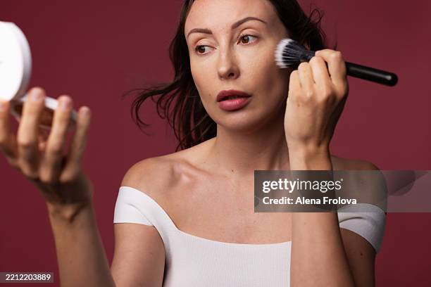 middle aged beautiful woman doing makeup holding brush in hands looking in mirrior on red background - hyperpigmentation stockfoto's en -beelden