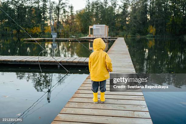 child wearing yellow raincoat and boots standing on wooden pier looking at lake - raincoat stock pictures, royalty-free photos & images