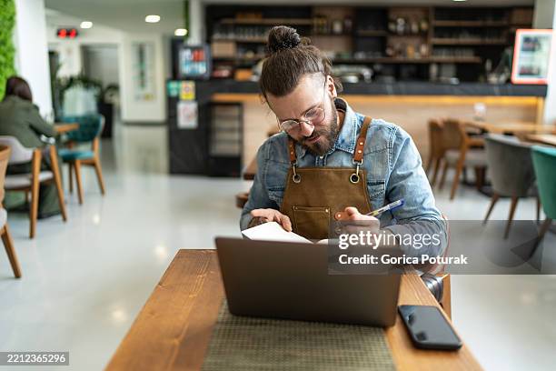coffee shop owner doing inventory using laptop and notebook - obter inventário imagens e fotografias de stock
