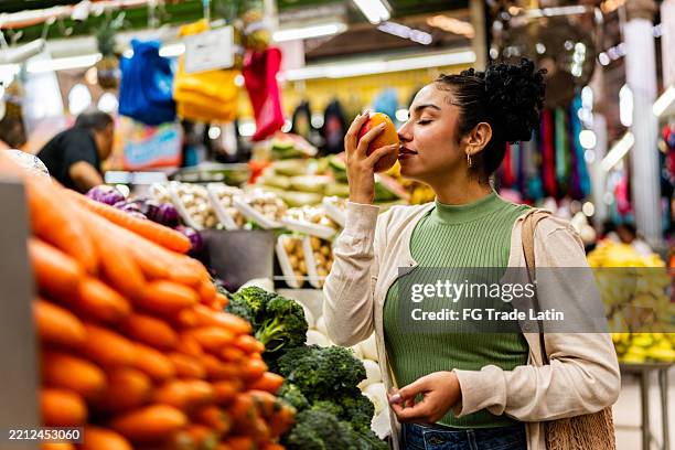 young woman buying fruits and smelling peach at market - mango fruta tropical fotografías e imágenes de stock