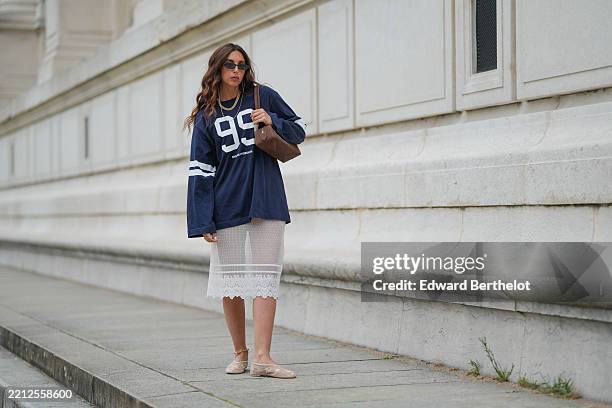 Gabriella Berdugo wears long, wavy brown hair with a center part, paired with a matte complexion, nude lipstick and natural brows. Black rectangular...