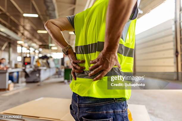 lavoratore in giubbotto ad alta visibilità che soffre di mal di schiena in ambiente industriale - ergonomia foto e immagini stock