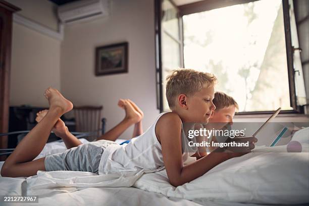 two little boys relaxing on a bed with tablets near an open window - digital native stock pictures, royalty-free photos & images