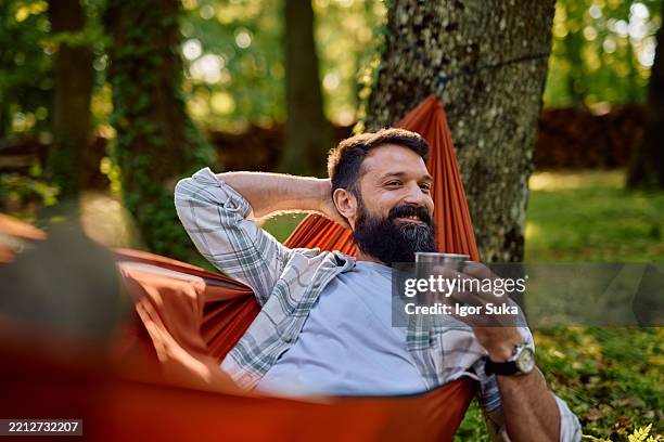 hombre barbudo relajándose en la hamaca bebiendo café en el bosque - hamaca fotografías e imágenes de stock